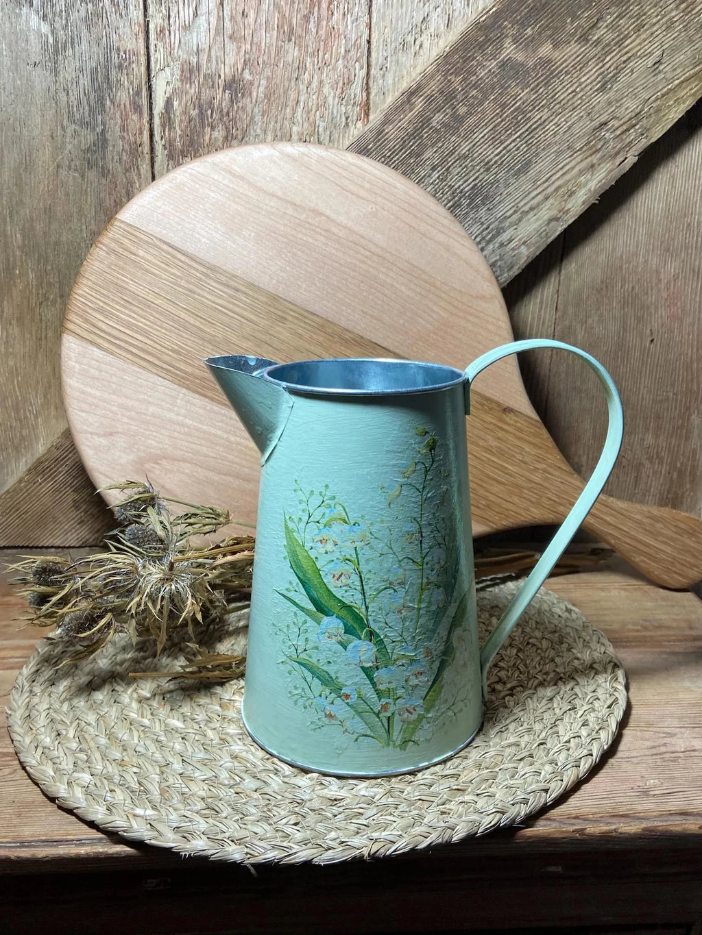 Teal metal pitcher with floral design on a woven mat against a wooden background